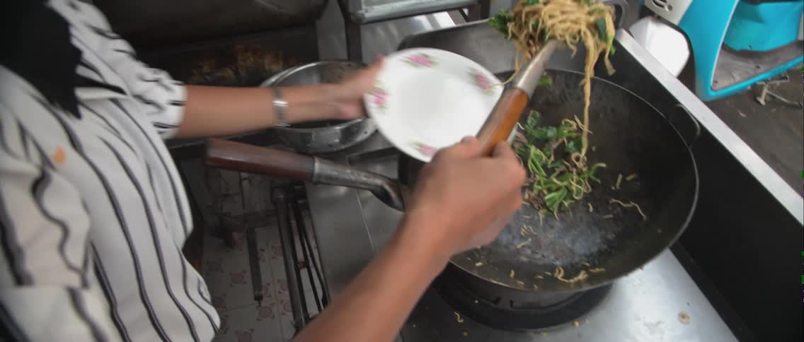 A woman cooks food in a large pan and scoops it onto a plate