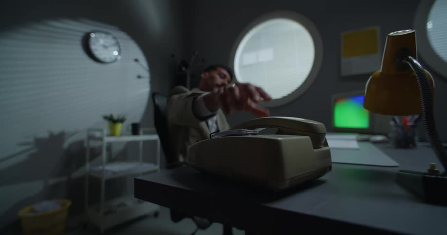A man sitting at his desk putting down an old style phone with an old computer on his desk