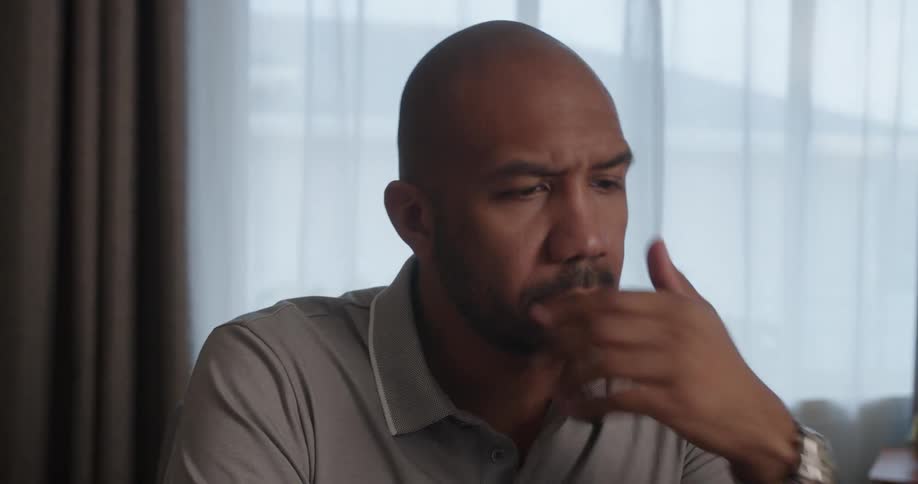 Bald man with gray shirt sits at desk looking at laptop in front of windows with curtains
