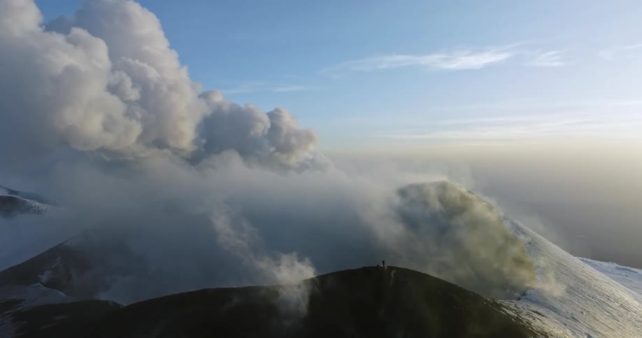 Aerial view of snow capped mountain with steam coming off clouds and man standing on peak