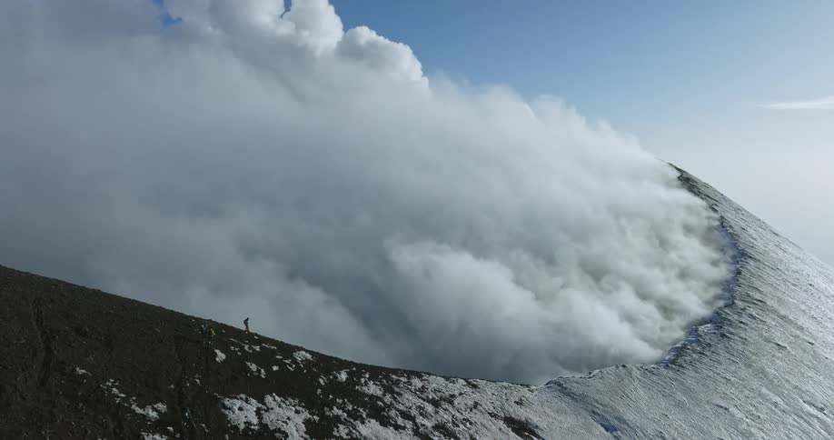 A time lapse of an aerial view of a steaming mountain with clouds rolling off it and a hiker on the ridge