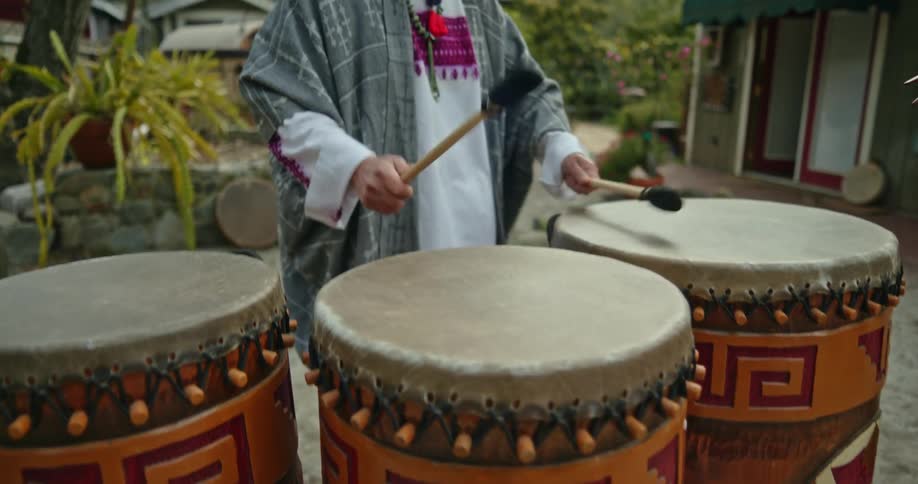 A native american man is standing outside of his house playing the drums and there are mountains in the distance