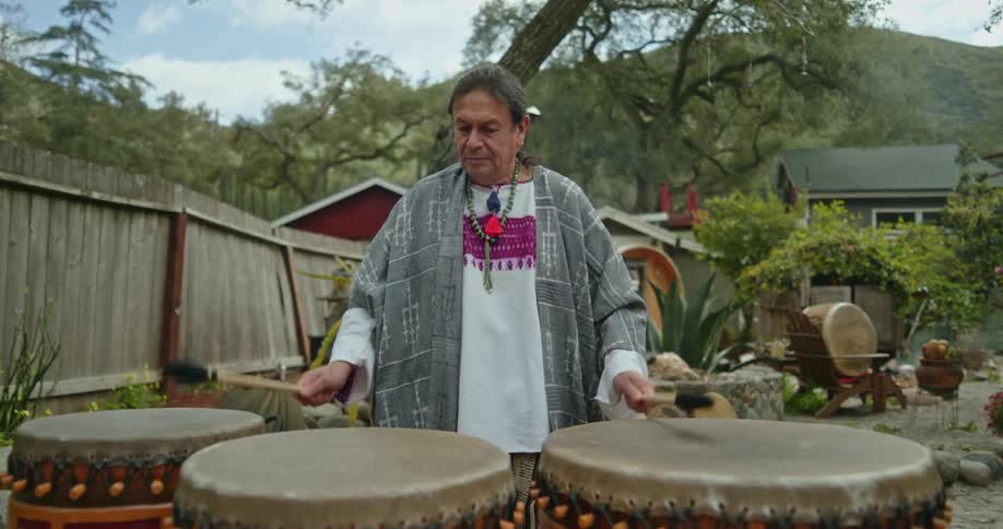 A native american man is standing outside of his house playing the drums and there is a mountain in the distance