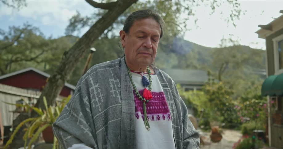 A native american man is standing outside near a building playing the drums and there are mountains in the distance