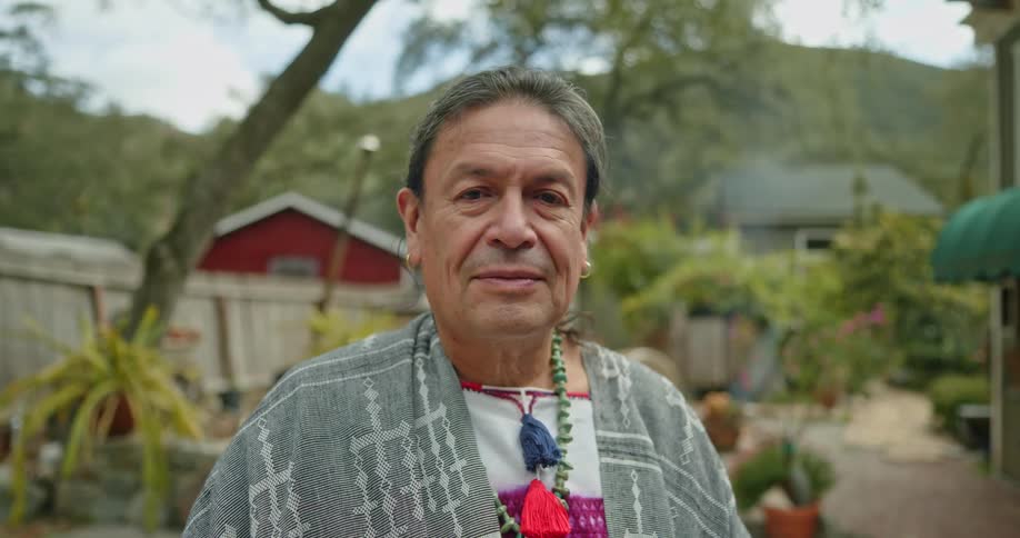 A native american man is standing outside near a building and there is a mountain in the distance