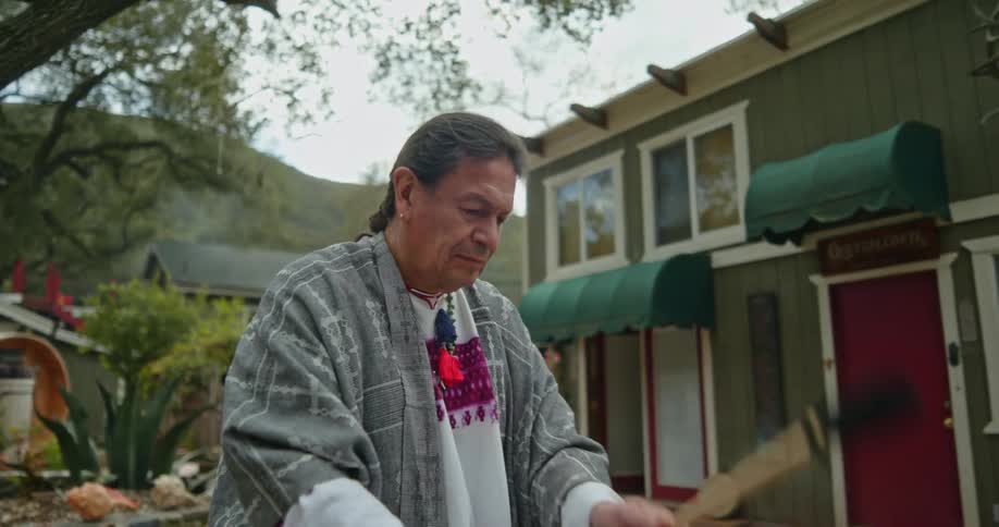 A native american man is standing outside of near a building playing the drums and there are mountains in the distance