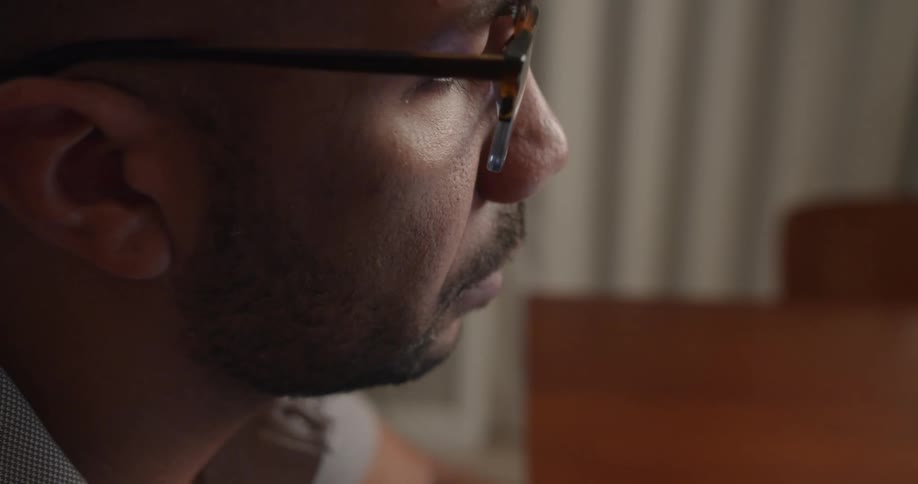 Bald man with glasses beard and gray shirt sits at desk working on laptop