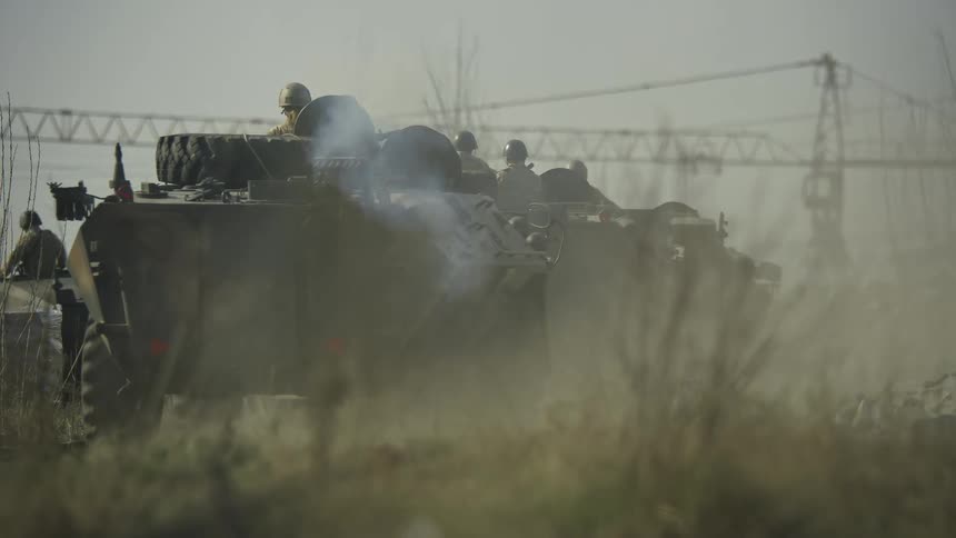A military caravan of trucks drives down a dusty rural road near power infrastructure
