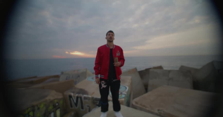 A close up of a man opening an aluminum can while standing on a large rock with the ocean in the background