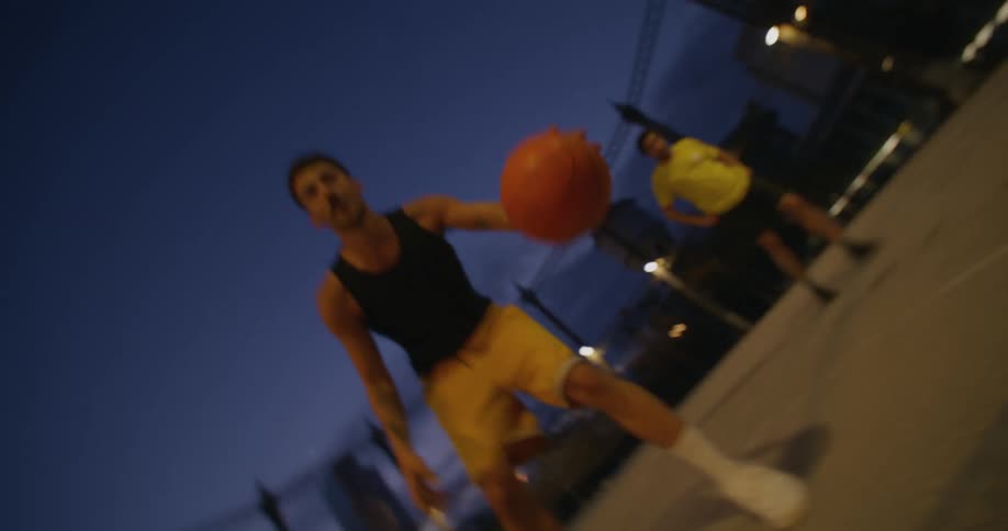 At night man dribbles basketball as another watches with skyscraper in the background