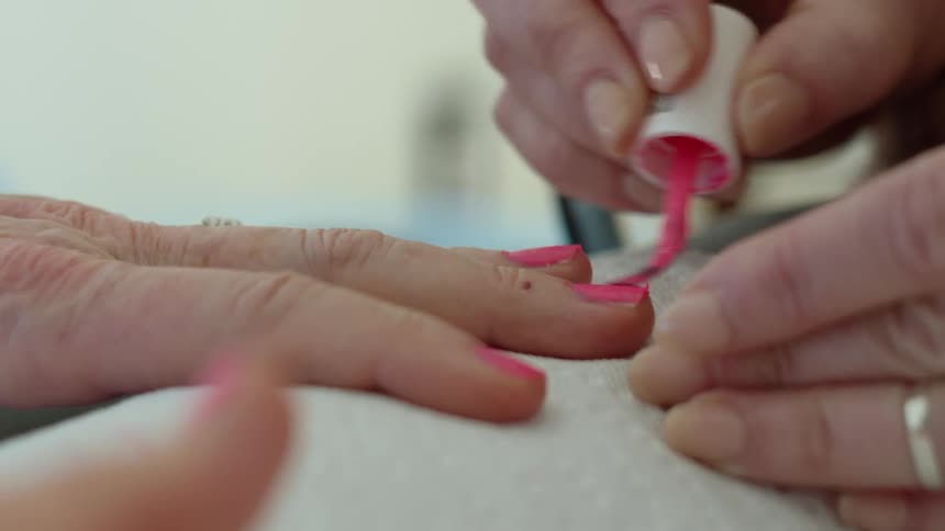 An extreme close up of a woman painting another womans nails with pink nail polish