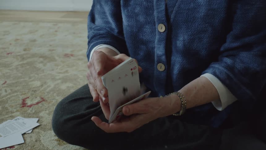 An elderly woman sits on a rug on the floor while shuffling a deck of cards