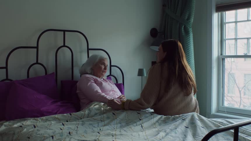 Woman sits on bed holding hand of older woman lying in bed