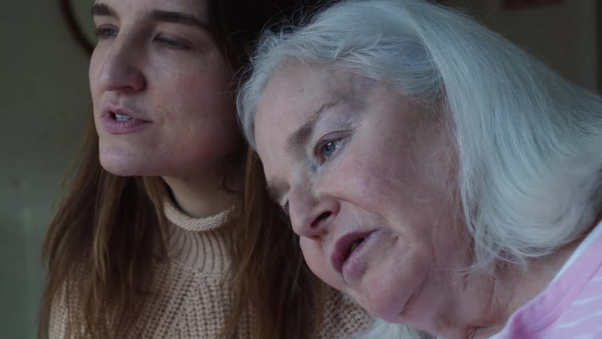 Young woman sitting and talking with grandmother while grandmother rests her head on granddaughters shoulder