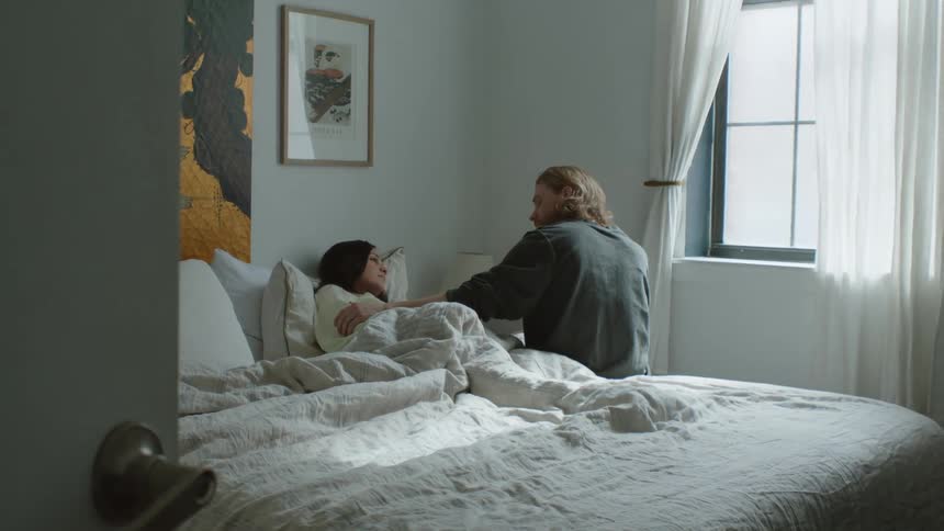 A man sits on the edge of a bed where a women lays in it under blankets looking sick