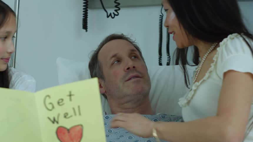 People visit man lying in hospital with a get well greeting card