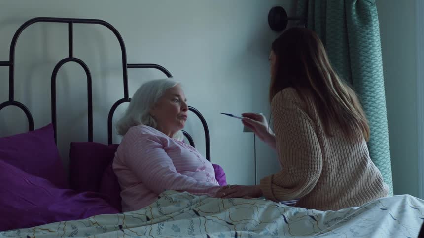 Woman takes temperature of elderly woman lying in bed and touches her forehead to feel temperature too