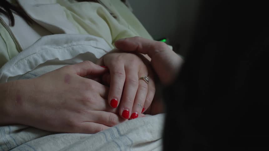 A males hand is shown stroking a females hand while she lays in bed