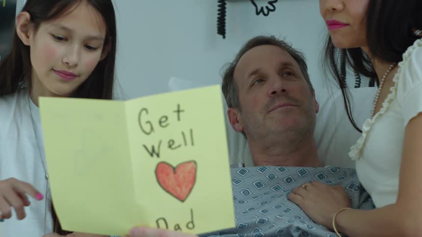 A daughter shares a get well card with her father in the hospital while her mother hugs him