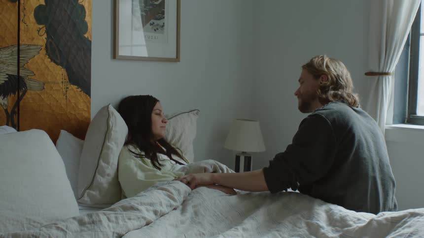 A woman laying in bed sick and sleeping as a man sits on the edge of the bed and kisses her on the forehead