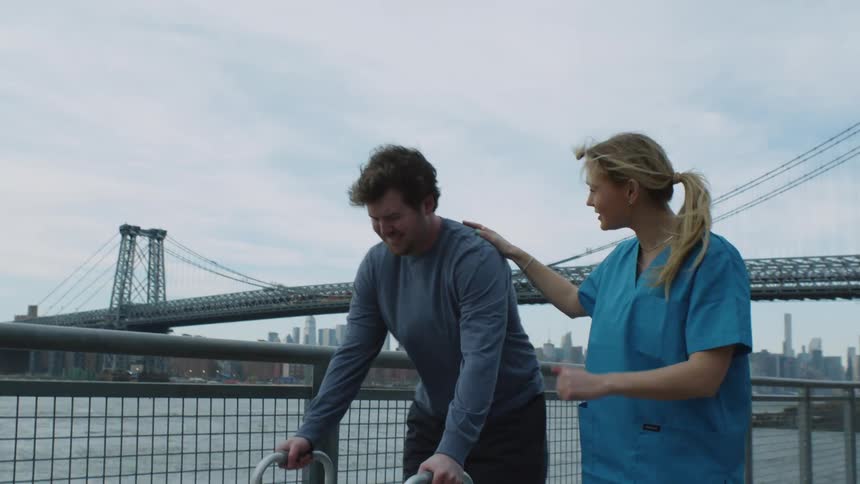 A woman nurse wearing blue walking beside a mal patient using a walker to walk slowly near a fence