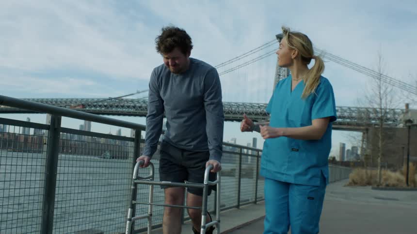 A nurse accompanies and encourages a man attempting to walk with a walker outdoors during the day