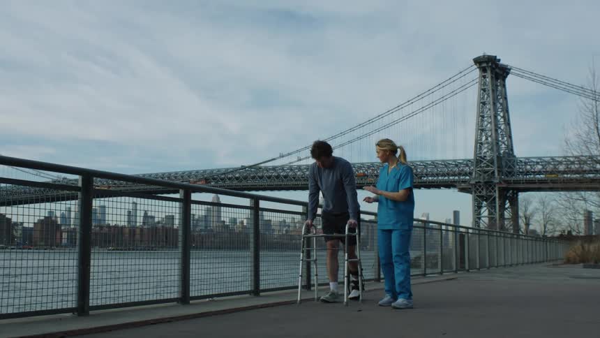 Medical professional claps while watching man wearing temp cast walk with a walker near water with suspension bridge and city in background