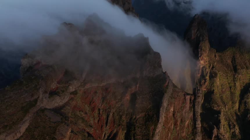 An aerial of a rocky mountain range with fog rolling out of the tops of the mountain