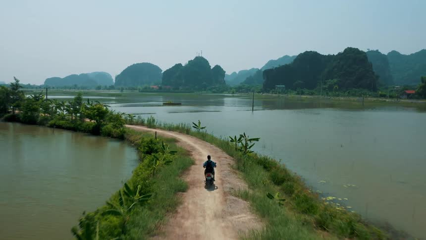 An adult rides a motorcycle down a dirt road in a lush tropical area with bodies of water located on each side of the roadway