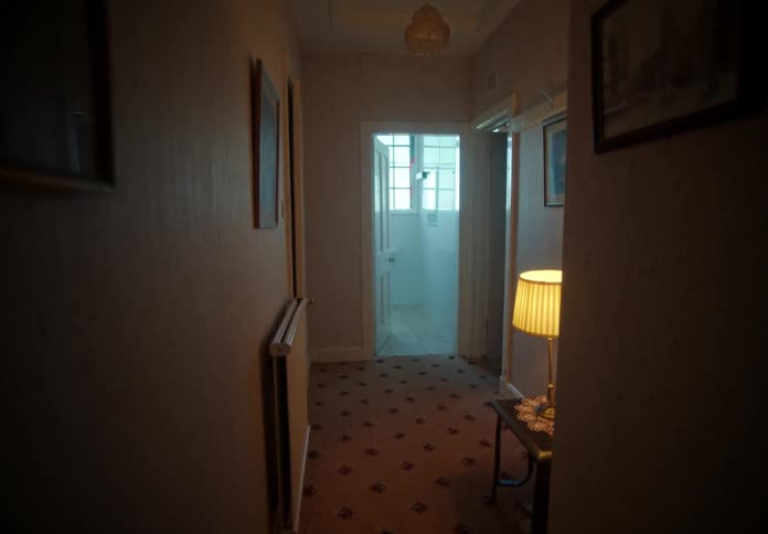 A table lamp is on in the hallway as light shines in a smoke filled room at the end of the hall