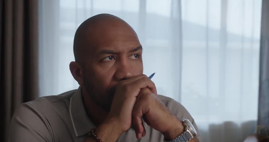 Bald man with gray shirt wrist watch and beaded wrist sits at table looking up and thinking