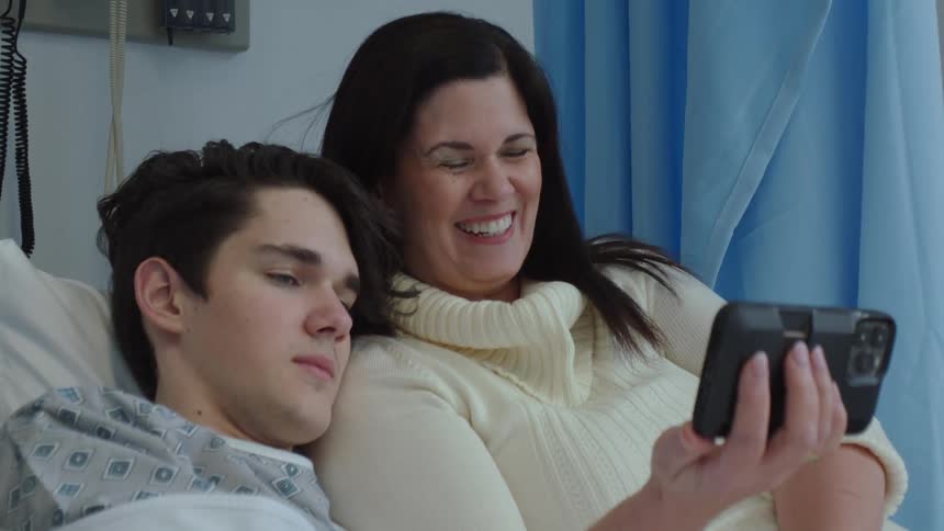 Mother visiting his son in the hospital rests with him on the bed while watching a video on the phone