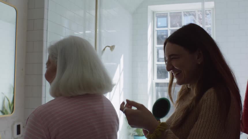 A grandaughter brushes her grandmothers hair as they laugh and talk in the bathroom