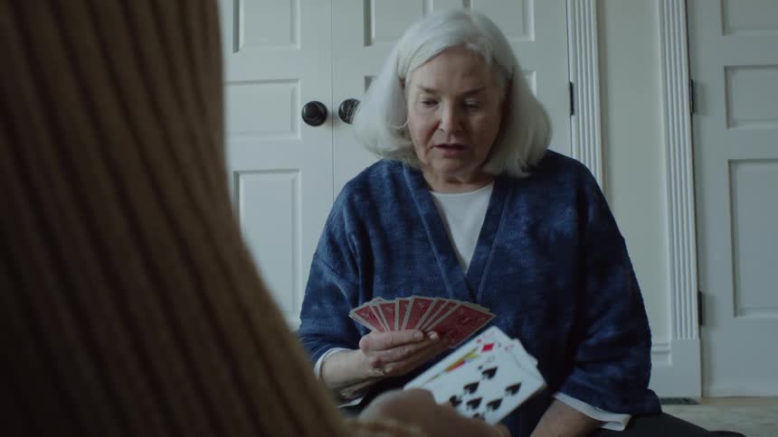 A close up of a persons arm who sits across from an older woman holding cards in her hand sitting on the floor