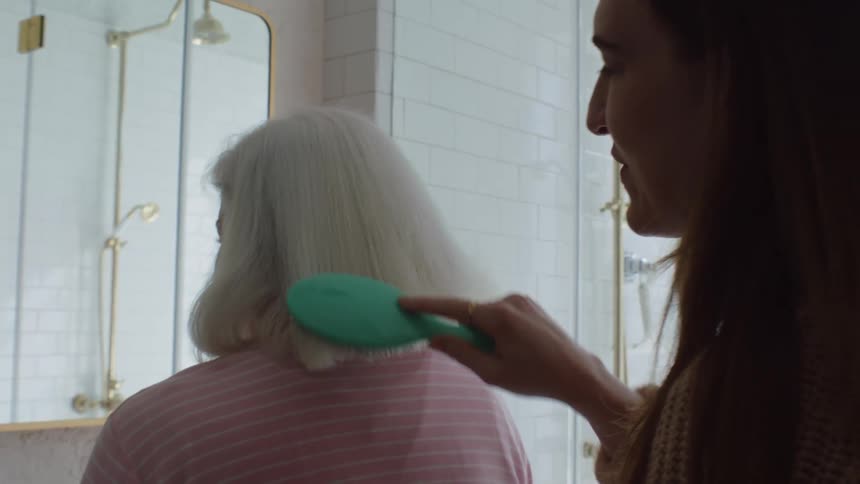 Young woman combs elderly womans hair in the bathroom laughing