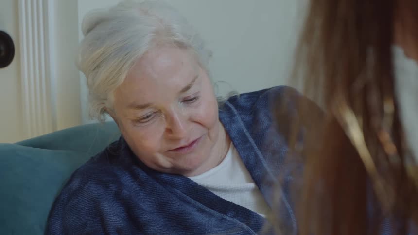 Close up of an elderly woman wearing a blue robe talking to a relative while resting
