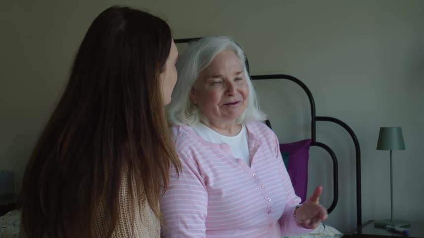 An elderly woman and a younger woman sitting on the edge of a bed talking laughing and smiling