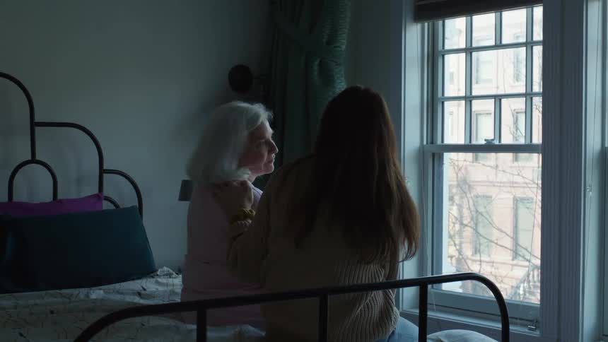 Young woman and grandmother sitting on bed in bedroom in front of window talking and bonding