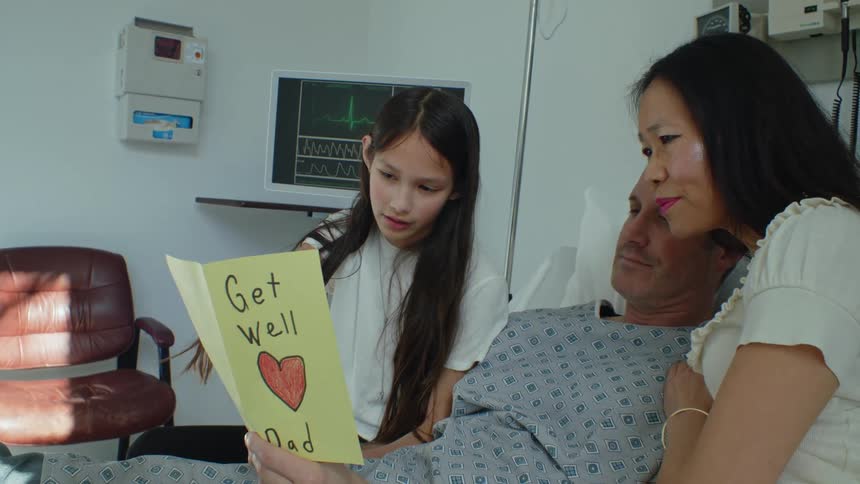 A father in a hospital bed reads a get well soon card with his wife and daughter