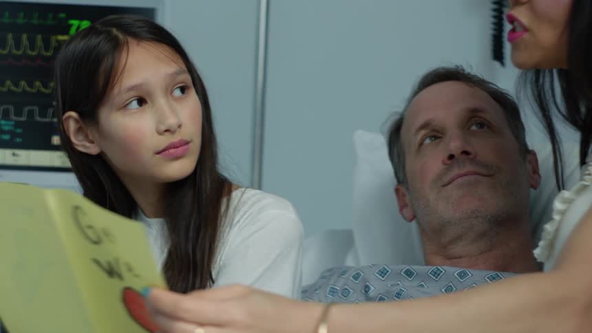 Man resting in a hospital bed reads a card on a visit from his wife and daughter