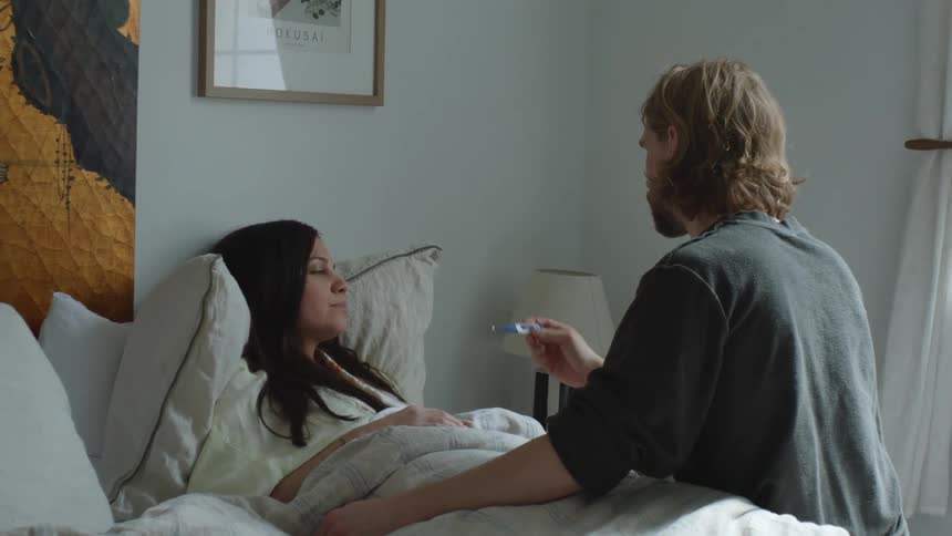A man lovingly takes his sick wifes temperature while she lays in bed at home