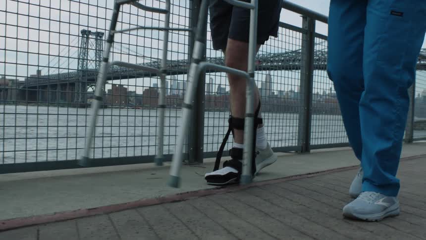 A nurse helps a man in a cast use a walker along a bridge