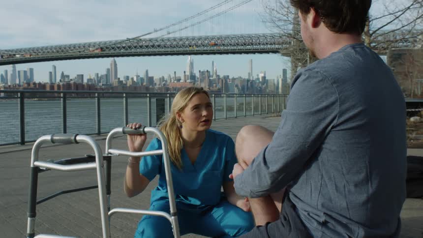 A man sitting down with his knee up as a nurse in blue kneels down in front of him and talks to him while holding his walker