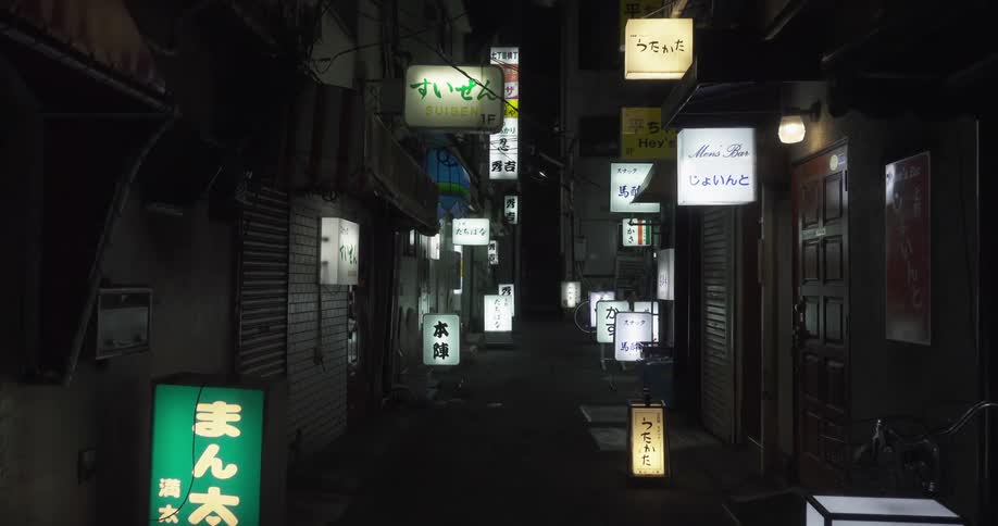 The camera moves down an alley in a japanese city lit up at night by electric signs