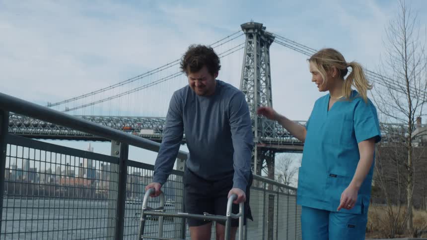 Female physical therapist or nurse walking alongside man with walker outside during the day in city