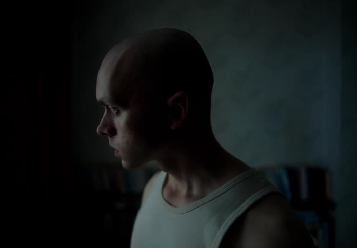 An up close of a man who is bald wearing a white tank top staring angrily in a dimly lit room
