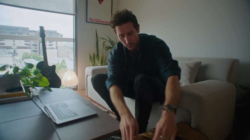 A man arrives home walks to his couch opens his laptop and takes a camera out of his bag
