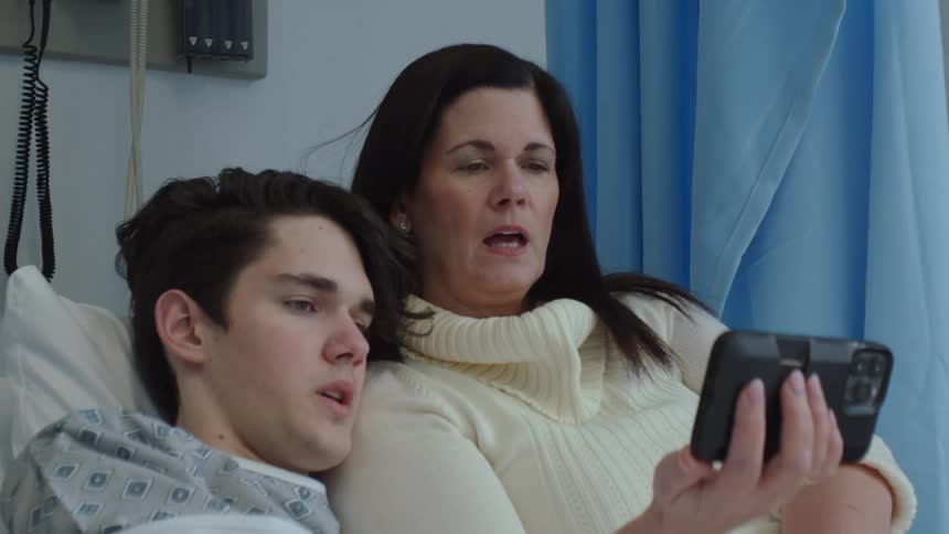 Mother sits with her son on a hospital bed watching a video on his phone