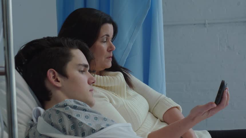A mother sits next to her teenage son in a hospital gown on a bed as she holds out her phone as he watches