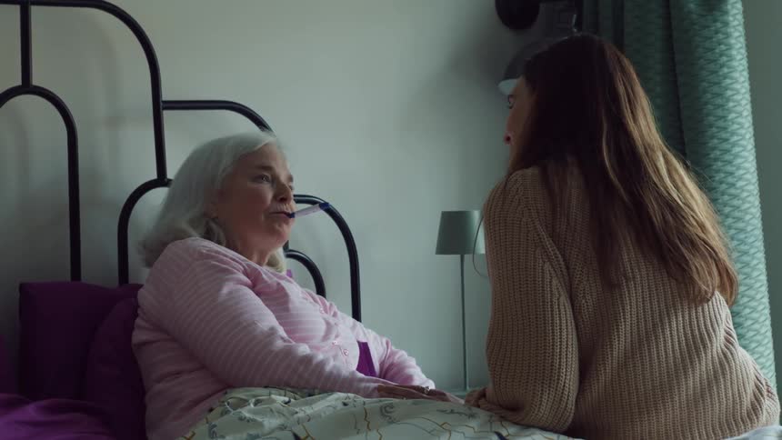 Young woman puts takes the temperature of an elderly woman resting in bed